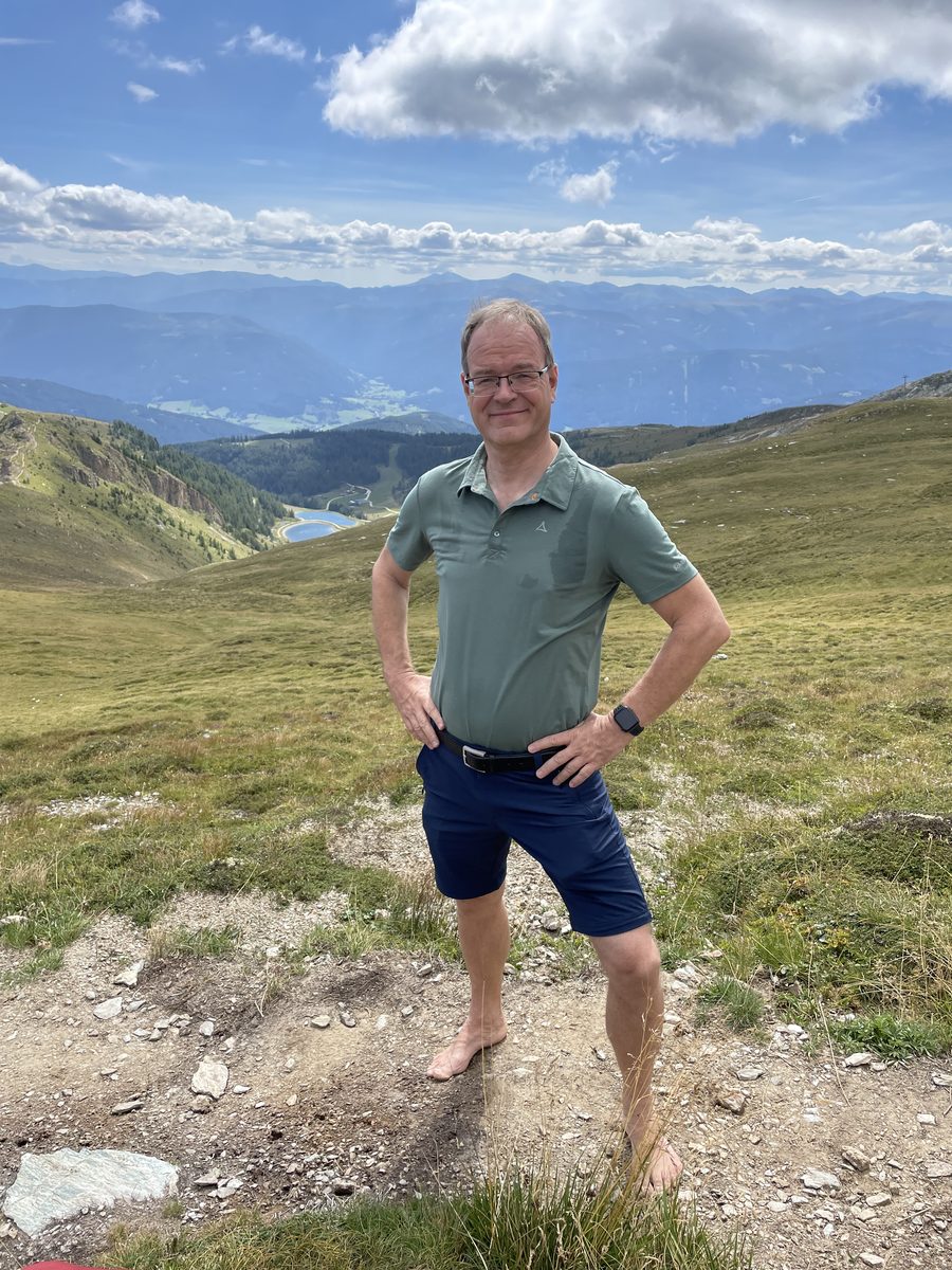 Nils steht barfuß auf einer Bergwiese mit Alpenpanorama im Hintergrund